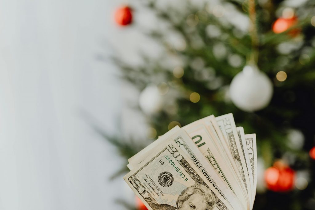 5 Tips to Balance Food and Gift Expenses This 2024 Holiday Season Stack of US dollar bills in front of a decorated Christmas tree, symbolizing holiday spending.